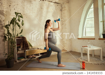 Determined young pregnant woman exercising with dumbbells at home Determined young pregnant woman exercising with dumbbells at home 141459333