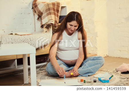 Focused young brunette pregnant woman painting belly while sitting on floor Focused young brunette pregnant woman painting belly while sitting on floor 141459339