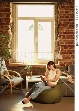 Pregnant woman reading book while relaxing on beanbag near window 141459340