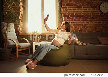 Pregnant woman relaxing on beanbag and using smartphone at home Pregnant woman relaxing on beanbag and using smartphone at home 141459342