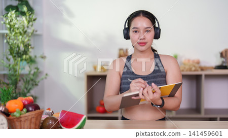 Healthy young woman wearing headphones and writing meal plan in kitchen 141459601