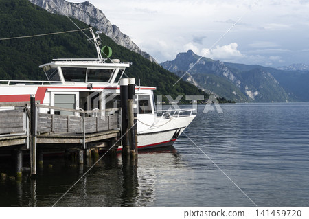 Passenger boat docked at a wooden pier on Lake Traunsee with an alpine mountain landscape, illustrating summer tourism in Austria. Passenger boat docked at a wooden pier on Lake Traunsee with an alpine mountain landscape, illustrating summer tourism in Austria. 141459720