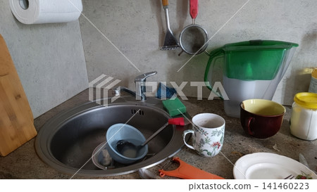Cluttered kitchen counter with a sink full of dirty dishes, sponges, utensils, mugs, a water filter pitcher, and hanging tools... Cluttered kitchen counter with a sink full of dirty dishes, sponges, utensils, mugs, a water filter pitcher, and hanging tools... 141460223