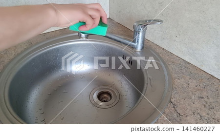 Close up of a hand using a bright green sponge to clean a wet stainless steel kitchen sink with visible drain and faucet.. Close up of a hand using a bright green sponge to clean a wet stainless steel kitchen sink with visible drain and faucet.. 141460227