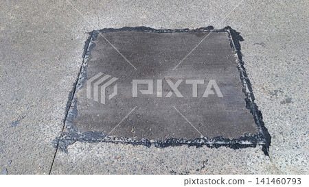 Rectangular fresh asphalt repair patch on light concrete background. Dark gray rough texture with wet sheen along edges. Natural daylight highlights contrast between old new pavement Rectangular fresh asphalt repair patch on light concrete background. Dark gray rough texture with wet sheen along edges. Natural daylight highlights contrast between old new pavement 141460793