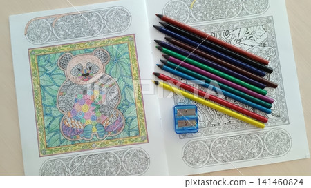Open coloring book with a panda mandala partially colored, colored pencils arranged diagonally across the right page, pencil sharpener included. Open coloring book with a panda mandala partially colored, colored pencils arranged diagonally across the right page, pencil sharpener included. 141460824
