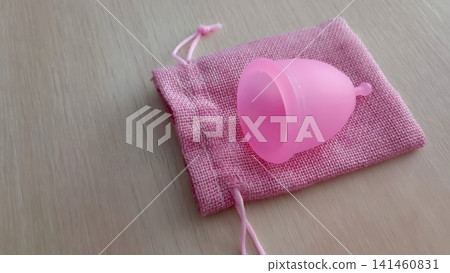 Bright pink menstrual cup sitting on a crumpled pink drawstring pouch on a light wooden surface, high angle close up. Bright pink menstrual cup sitting on a crumpled pink drawstring pouch on a light wooden surface, high angle close up. 141460831