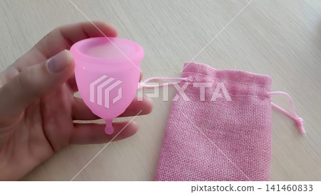 Hand presenting a pink menstrual cup next to a woven pink drawstring pouch on a light wooden surface. Hand presenting a pink menstrual cup next to a woven pink drawstring pouch on a light wooden surface. 141460833