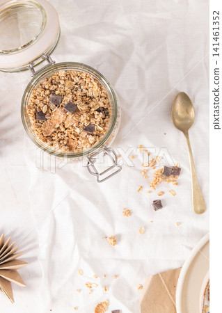 Crunchy granola with dark chocolate chunks in a glass jar set on a white fabric background for a healthy and nutritious morning breakfast meal Crunchy granola with dark chocolate chunks in a glass jar set on a white fabric background for a healthy and nutritious morning breakfast meal 141461352