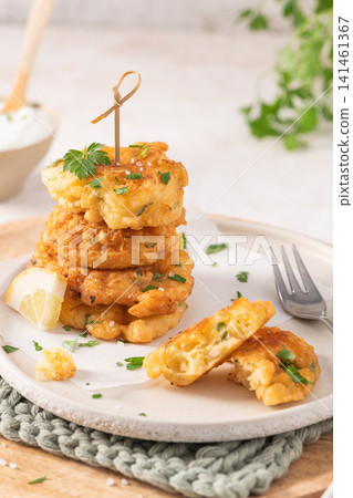 A stack of golden cod fritters on a textured plate with fresh parsley, lemon, and a fork, offering a savory Mediterranean-style seafood appetizer snack A stack of golden cod fritters on a textured plate with fresh parsley, lemon, and a fork, offering a savory Mediterranean-style seafood appetizer snack 141461367