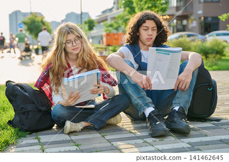 Two teenage students sitting outdoors reading workbook preparing for exams together Two teenage students sitting outdoors reading workbook preparing for exams together 141462645