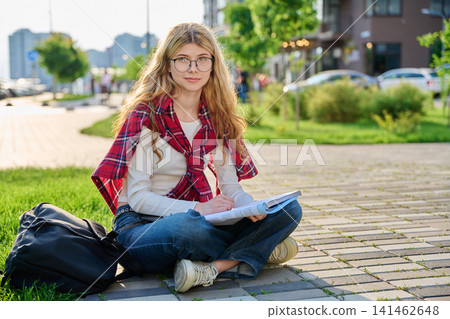 Happy young female student sitting on ground with notebook in city street 141462648