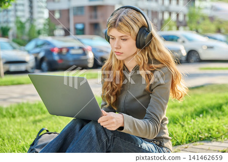 Young woman in headphones working on laptop outdoors sitting on green grass 141462659