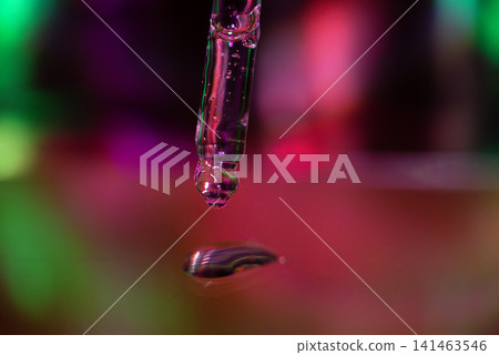 Close-up of a dropper releasing a drop of purple liquid onto a reflective surface, with colorful bokeh lights creating a vibrant background Close-up of a dropper releasing a drop of purple liquid onto a reflective surface, with colorful bokeh lights creating a vibrant background 141463546