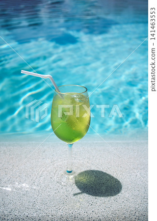 Green cocktail in a clear glass with a straw placed on the edge of a swimming pool, with shimmering blue water reflecting sunlight in the background Green cocktail in a clear glass with a straw placed on the edge of a swimming pool, with shimmering blue water reflecting sunlight in the background 141463553