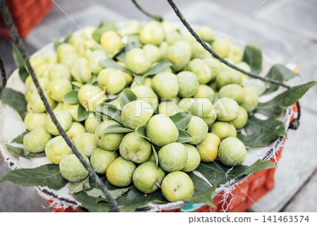 Fresh green citrus fruits arranged in a woven basket with leaves, suspended by ropes, placed on a textured surface in a market setting 141463574