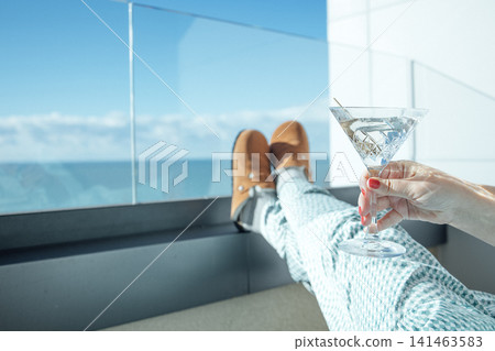 Woman in patterned pants and brown shoes relaxes by a glass railing holding a martini glass with ocean view in the background under a clear blue sky Woman in patterned pants and brown shoes relaxes by a glass railing holding a martini glass with ocean view in the background under a clear blue sky 141463583