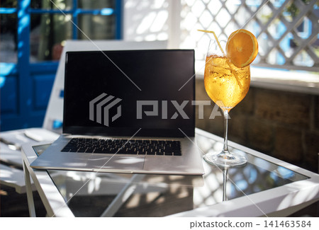 Laptop computer with blank screen beside a refreshing orange cocktail on a glass table in a bright outdoor setting with blue door and lattice background Laptop computer with blank screen beside a refreshing orange cocktail on a glass table in a bright outdoor setting with blue door and lattice background 141463584