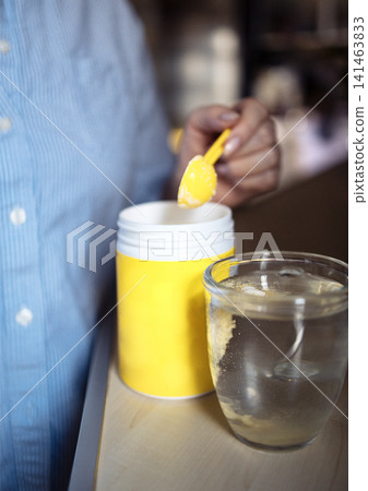 Person in blue shirt using yellow spoon to scoop powder from yellow container into clear glass of water on wooden surface in a kitchen setting 141463833