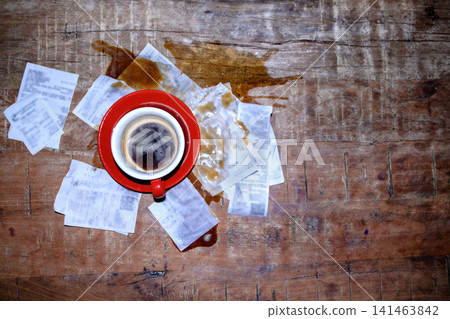 Red coffee cup filled with black coffee placed on a wooden table surrounded by spilled coffee and crumpled receipts scattered around 141463842
