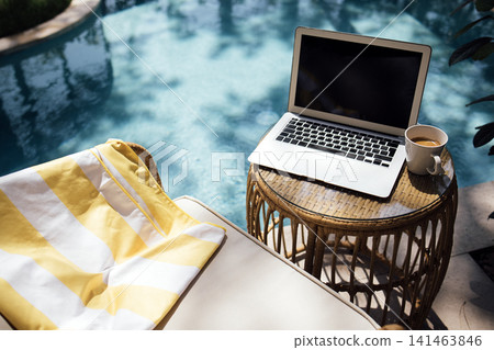 Laptop on a round wicker table beside a swimming pool, with a yellow and white striped towel draped over a lounge chair and a cup of coffee nearby Laptop on a round wicker table beside a swimming pool, with a yellow and white striped towel draped over a lounge chair and a cup of coffee nearby 141463846