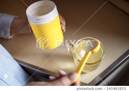 Person holding yellow container of powdered supplement while preparing drink in clear glass cup on wooden kitchen countertop with soft lighting Person holding yellow container of powdered supplement while preparing drink in clear glass cup on wooden kitchen countertop with soft lighting 141463853
