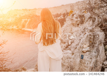Woman cliff sunset standing on a rocky coastal ledge looking at the ocean with long flowing hair during golden hour Woman cliff sunset standing on a rocky coastal ledge looking at the ocean with long flowing hair during golden hour 141464246
