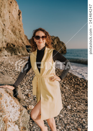Woman beach fashion stylish lady in yellow vest and sunglasses stands on a rocky shore with sea background Woman beach fashion stylish lady in yellow vest and sunglasses stands on a rocky shore with sea background 141464247