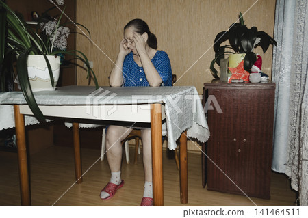 elderly woman blue top sits table hand resting against face she appears unwell quiet domestic scene conveys vulnerability, fatigue, need for care set against familiar backdrop simple home interior elderly woman blue top sits table hand resting against face she appears unwell quiet domestic scene conveys vulnerability, fatigue, need for care set against familiar backdrop simple home interior 141464511