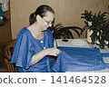 An elderly woman in blue dress focuses intently sewing garment table quiet domestic scene highlighting craftsmanship, patience, and the timeless joy creating clothing hand cozy familiar home setting 141464528