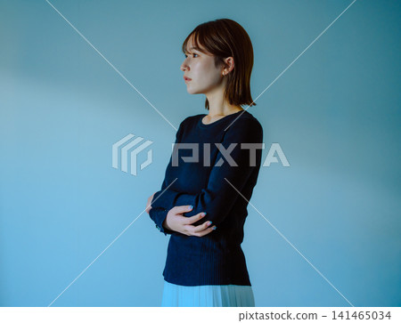 A woman with her arms crossed, looking to the side, against a blue background. 141465034