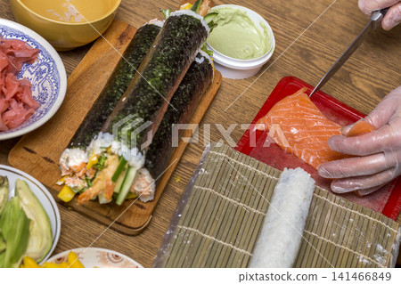 Making sushi and rolls at home. Plates with ingredients for traditional Japanese food and sushi rolls on wooden board on kitchen table, view from above. Making sushi and rolls at home. Plates with ingredients for traditional Japanese food and sushi rolls on wooden board on kitchen table, view from above. 141466849