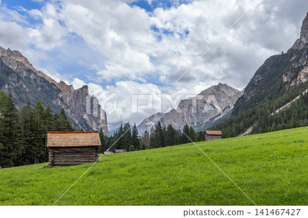 Wooden alpine huts in a green Val dai Tamersc meadow below Dolomite peaks Wooden alpine huts in a green Val dai Tamersc meadow below Dolomite peaks 141467427