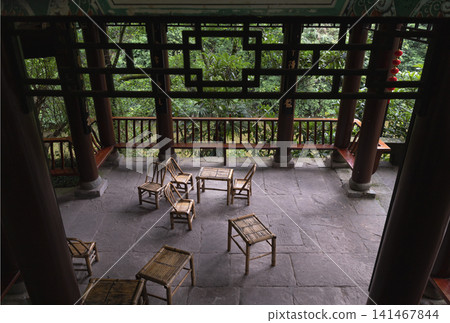 A serene high-angle perspective looking into a traditional open-air Chinese wooden pavilion furnished with handcrafted bamboo chairs and tables. 141467844