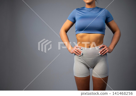 Fit woman in blue crop top and gray athletic shorts standing with hands on hips Fit woman in blue crop top and gray athletic shorts standing with hands on hips 141468256