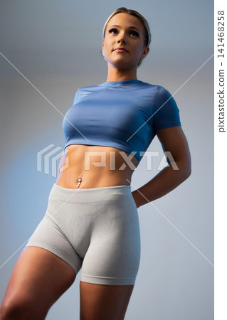 Fit woman in blue crop top and gray leggings posing confidently against a soft blue background Fit woman in blue crop top and gray leggings posing confidently against a soft blue background 141468258