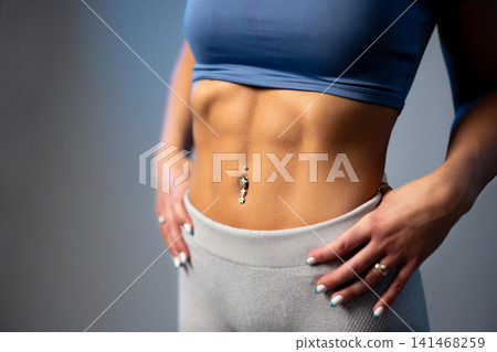 Fit woman showing defined abs in blue sports top and gray leggings with belly button piercing Fit woman showing defined abs in blue sports top and gray leggings with belly button piercing 141468259