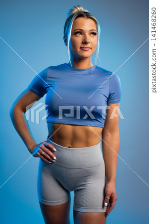 Fit young woman in blue crop top and gray leggings posing confidently against blue background Fit young woman in blue crop top and gray leggings posing confidently against blue background 141468260