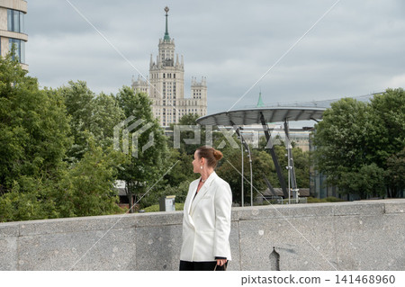 Woman stands in Zaryadye Park with Kotelnicheskaya Embankment Building and modern park architecture in background on cloudy day. Urban lifestyle, travel, sightseeing, city landmark, outdoor recreation 141468960
