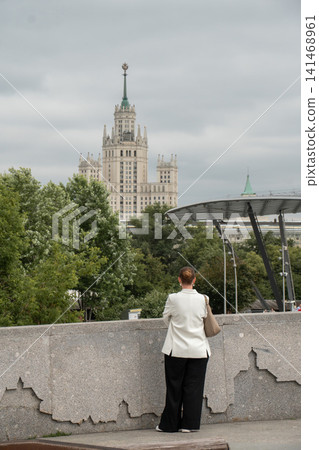 Woman stands in Zaryadye Park with Kotelnicheskaya Embankment Building and modern park architecture in background on cloudy day. Urban lifestyle, travel, sightseeing, city landmark, outdoor recreation Woman stands in Zaryadye Park with Kotelnicheskaya Embankment Building and modern park architecture in background on cloudy day. Urban lifestyle, travel, sightseeing, city landmark, outdoor recreation 141468961
