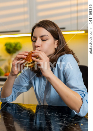 Young woman enjoying tasty burger in home kitchen indoors satisfied with quick fast food cheat lunch Young woman enjoying tasty burger in home kitchen indoors satisfied with quick fast food cheat lunch 141469386