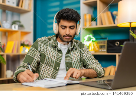Indian man on home table taking notes in headphones during online lesson with serious focused face Indian man on home table taking notes in headphones during online lesson with serious focused face 141469390