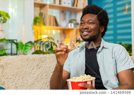 African American young man on home sofa watching film with popcorn and smiling in cozy living room African American young man on home sofa watching film with popcorn and smiling in cozy living room 141469399
