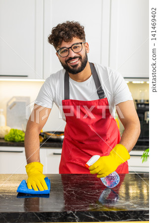 Arabian young adult man cleaning kitchen counter using spray bottle and microfiber cloth for hygiene 141469402