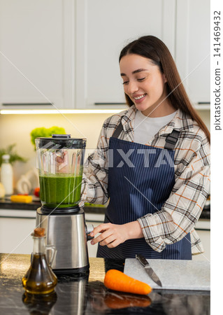 Young woman starts blender for green smoothie with spinach, banana, apple and carrot in kitchen 141469432