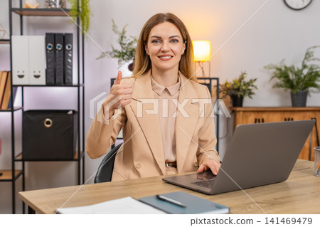 Businesswoman works on laptop shows thumbs up like gesture smiles approves positive results proud up Businesswoman works on laptop shows thumbs up like gesture smiles approves positive results proud up 141469479