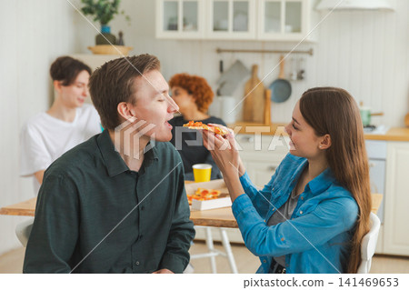 Home party. Girl feeding guy with piece of pizza. Friends spending time together having fun laughing at home. Happy diverse group eating pizza. Mixed race young buddies best friends enjoying weekend Home party. Girl feeding guy with piece of pizza. Friends spending time together having fun laughing at home. Happy diverse group eating pizza. Mixed race young buddies best friends enjoying weekend 141469653