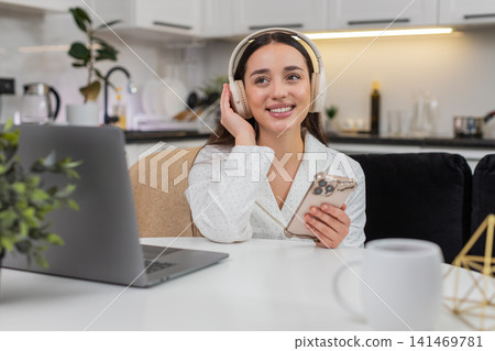Young woman wears wireless headphones types message on smartphone takes break with laptop nearby 141469781