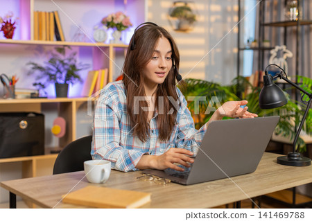 Young woman on home table using headset laptop for calm customer support video call in living room Young woman on home table using headset laptop for calm customer support video call in living room 141469788