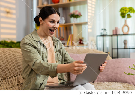 Young woman on home sofa celebrating success after good news on laptop with happy surprise indoors Young woman on home sofa celebrating success after good news on laptop with happy surprise indoors 141469790